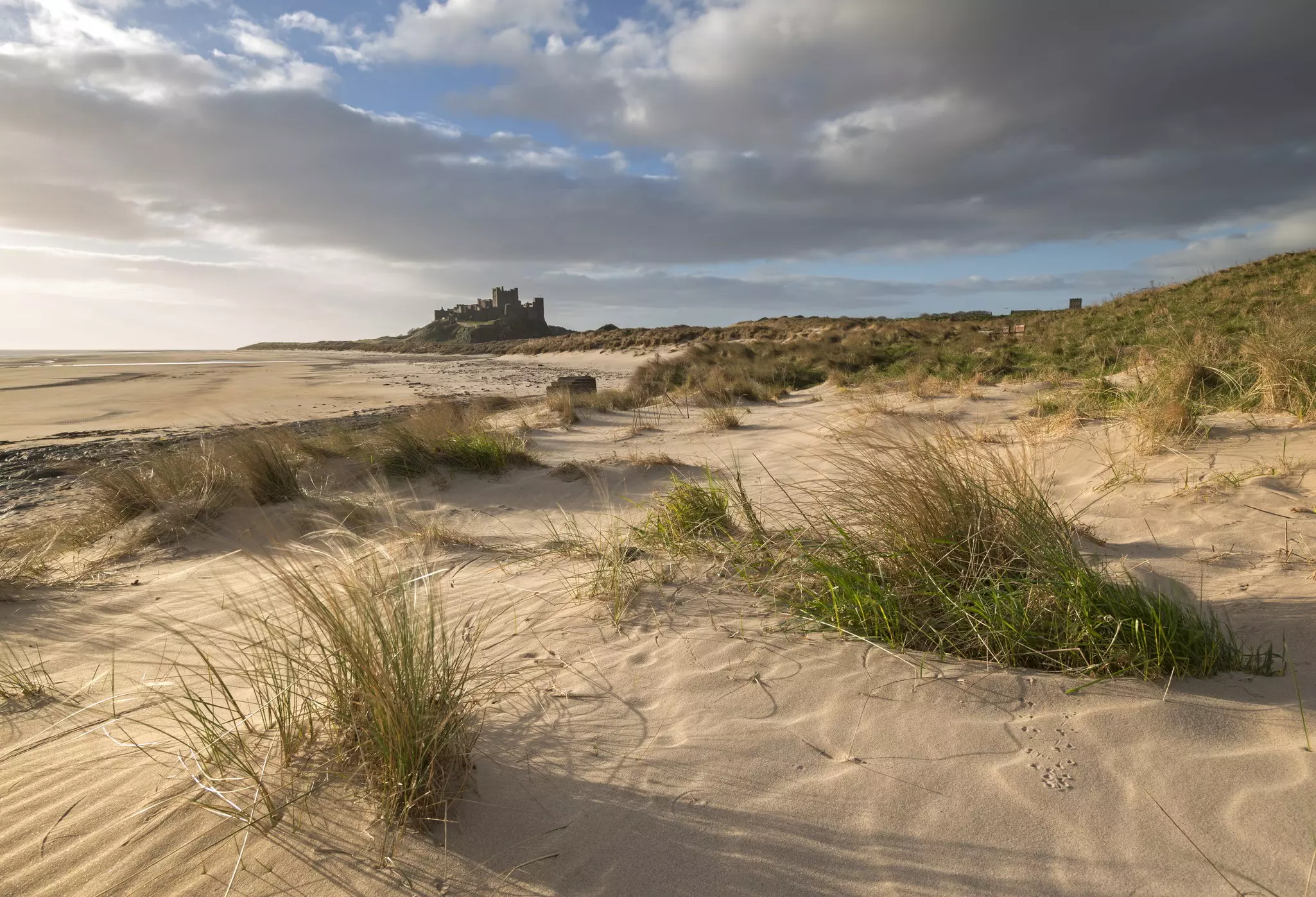 Grass topped sand dunes lead to a wide open beach. In the distance a medieval castle sits on a bluff.
