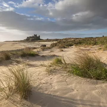 Grass topped sand dunes lead to a wide open beach. In the distance a medieval castle sits on a bluff.