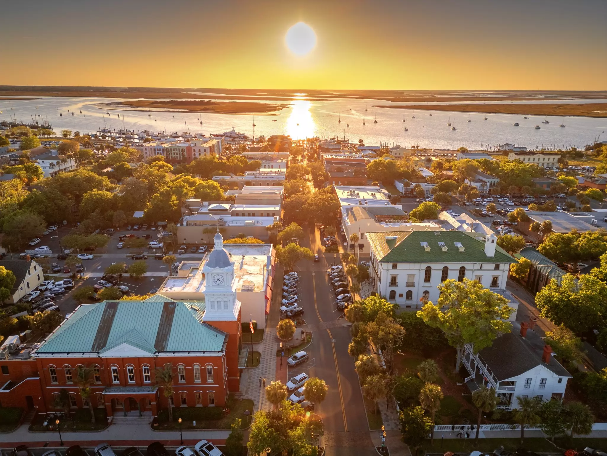 An aerial view of buildings in a village illuminated by the setting sun. Sailboats bob in an inlet in the distance.