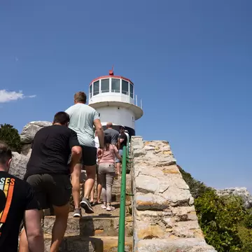 People walking up a stone stairway with a green handrail towards a white lighthouse under a blue sky.
