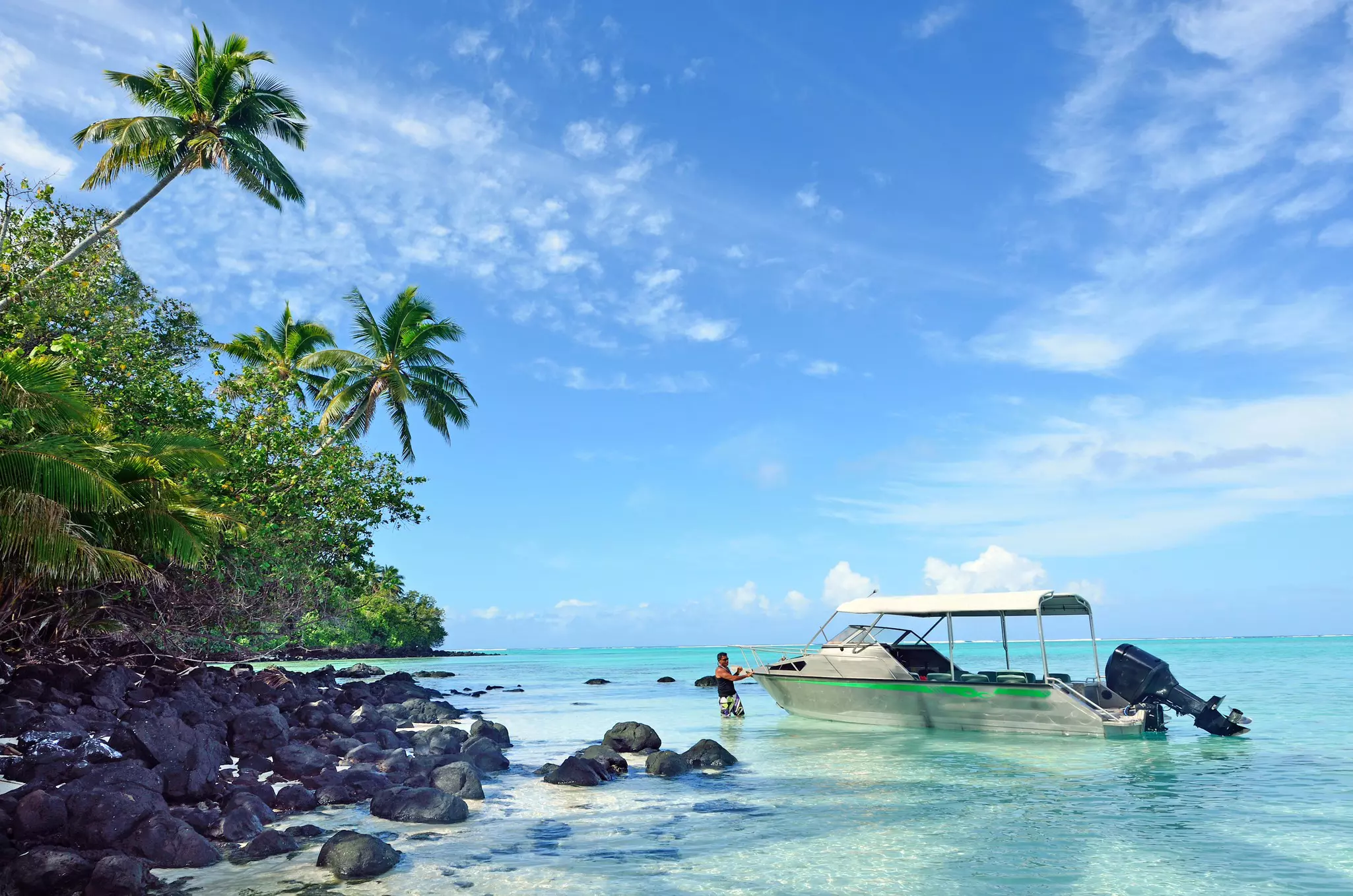 A man stands in shallow water near a moored motorboat off the shore of a tropical island.