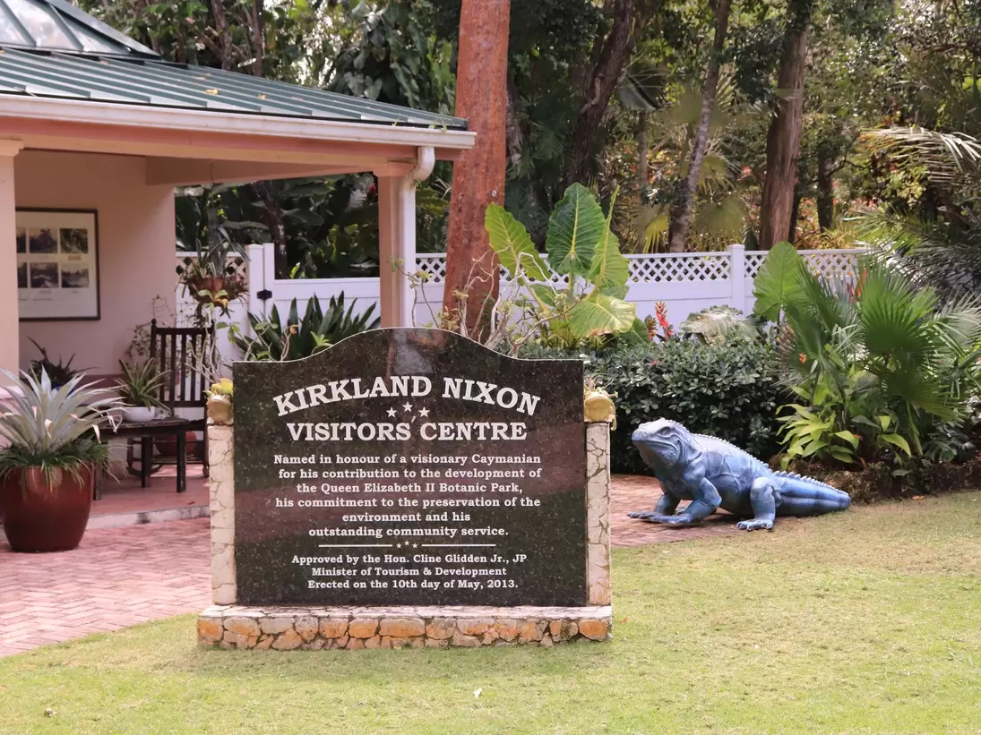 Stone sign for the Kirkland Nixon Visitors Centre beside a blue iguana sculpture, tropical plants and a veranda