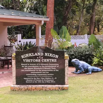Stone sign for the Kirkland Nixon Visitors Centre beside a blue iguana sculpture, tropical plants and a veranda