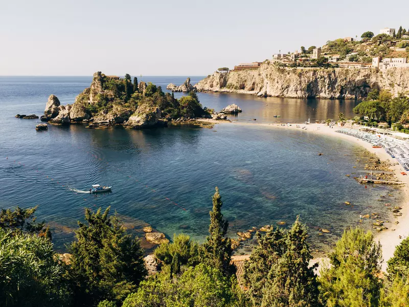 A boat in a blue cove in Italy; there is a sandy beach and a large rock formation in the water.