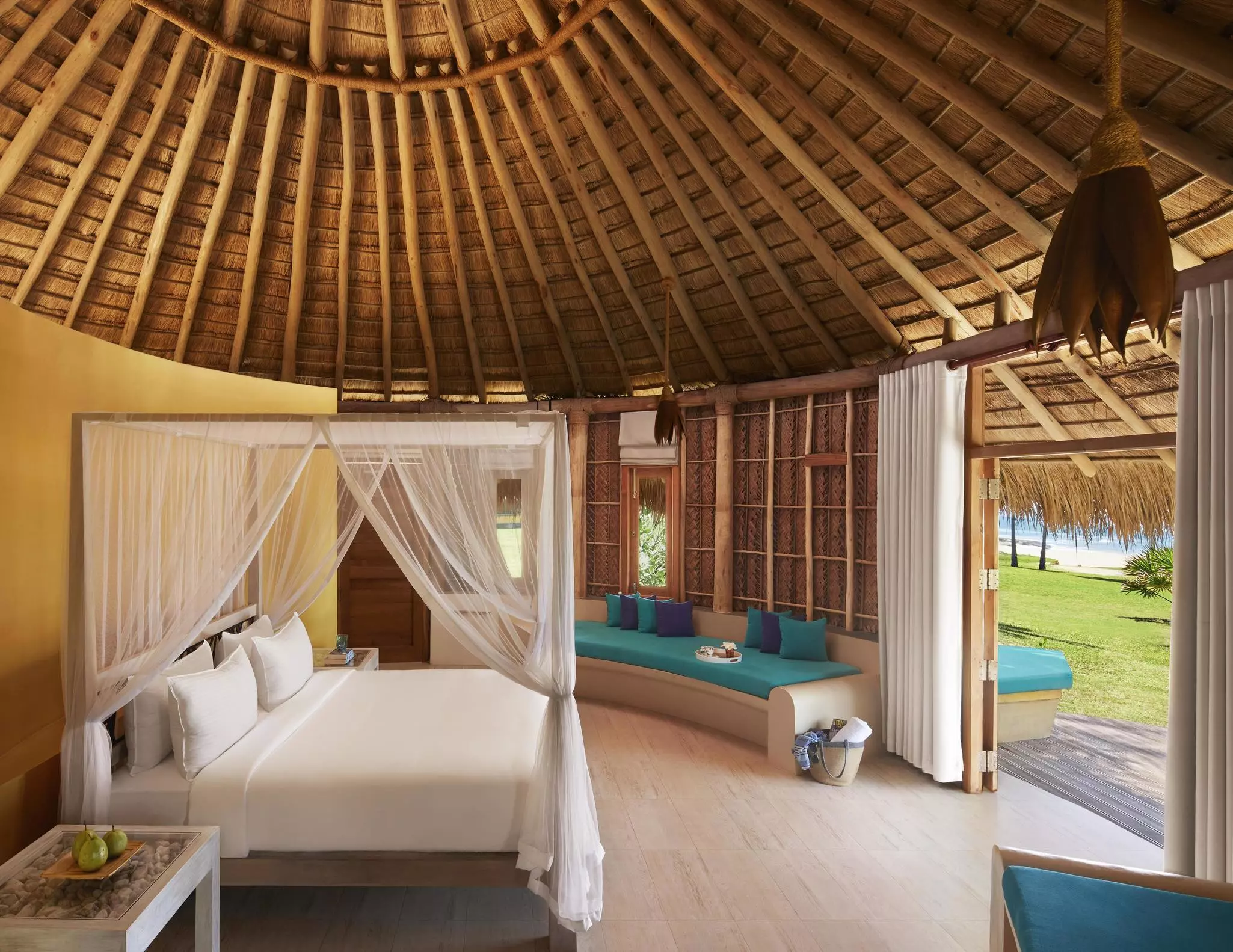 A large double bed in a thatched luxury bungalow with the edge of a sandy beach and the ocean outside.