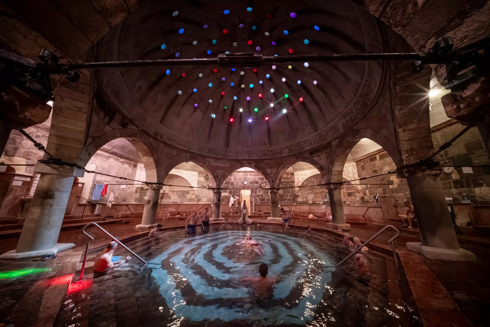 A wide view of a thermal bath surrounded by large columns and pointed arches, under a dome with small openings letting in light.