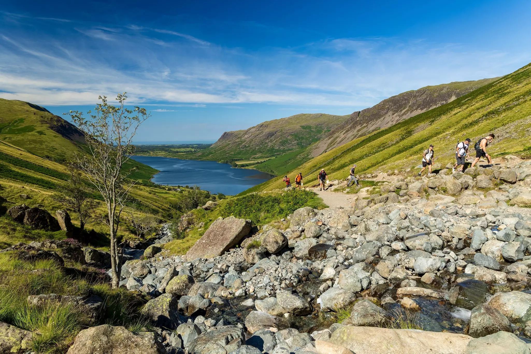Hikers follow a path uphill with a large lake behind them.
