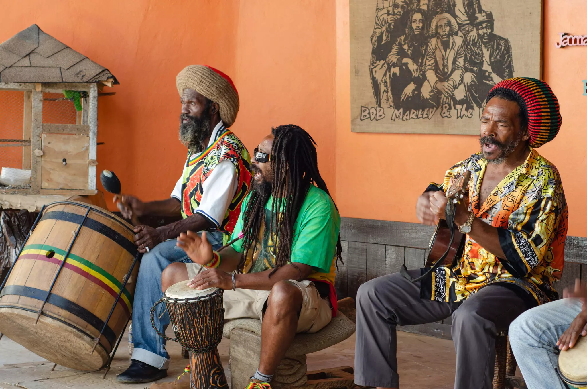 Three reggae musicians perform while seated against an apricot-colored wall.