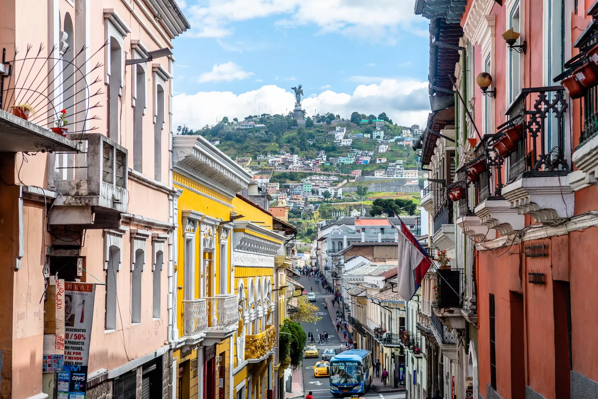 The facades of historic buildings line a narrow street in a city. A hill topped by a statue is seen in the distance.