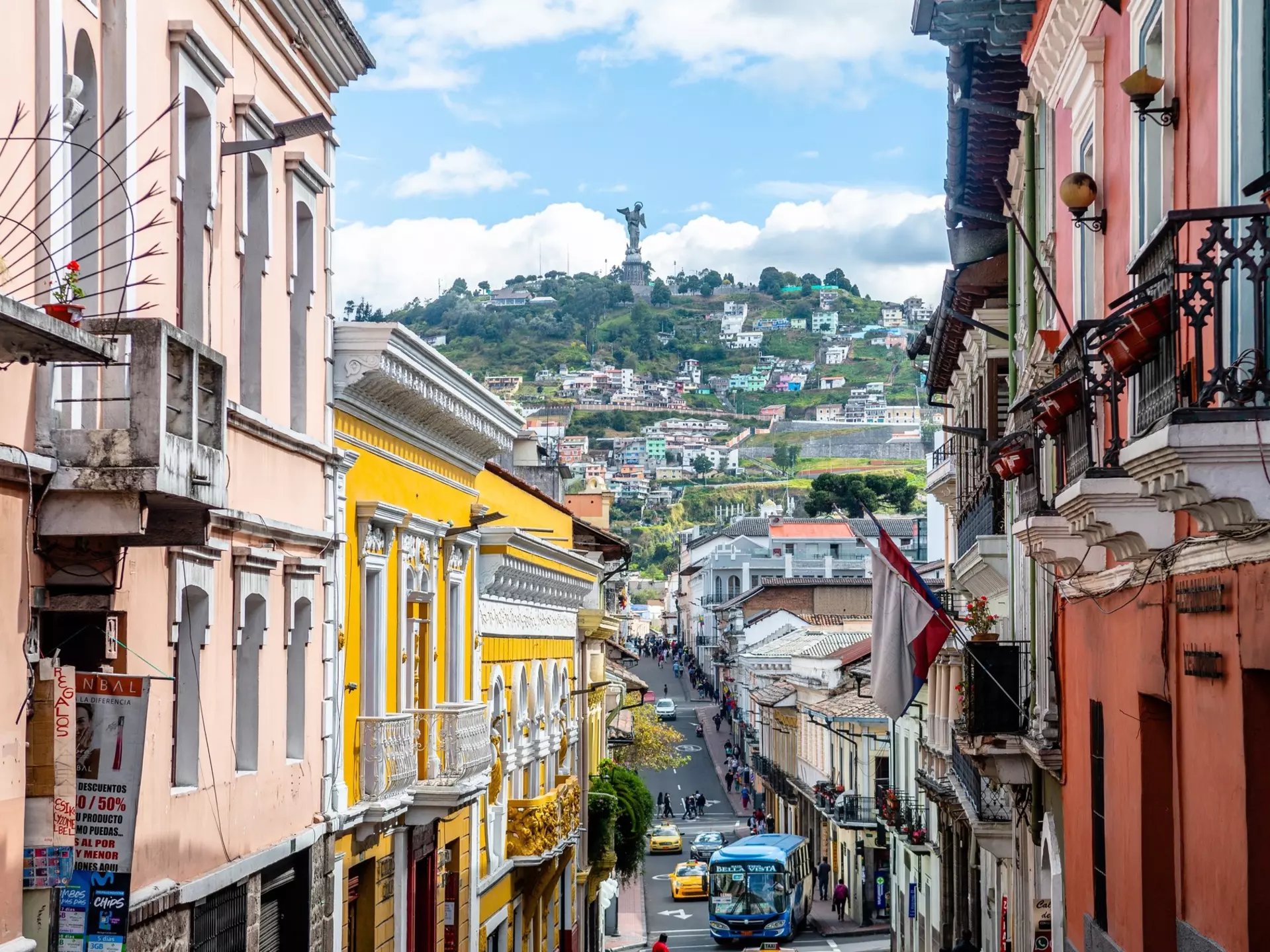 The facades of historic buildings line a narrow street in a city. A hill topped by a statue is seen in the distance.