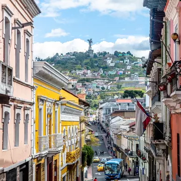 The facades of historic buildings line a narrow street in a city. A hill topped by a statue is seen in the distance.