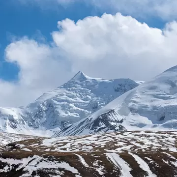Snow-covered mountain peaks under billowing white clouds, with a brown ridge streaked with snow in the foreground.