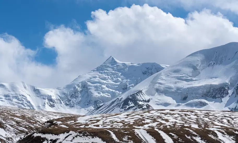 Snow-covered mountain peaks under billowing white clouds, with a brown ridge streaked with snow in the foreground.