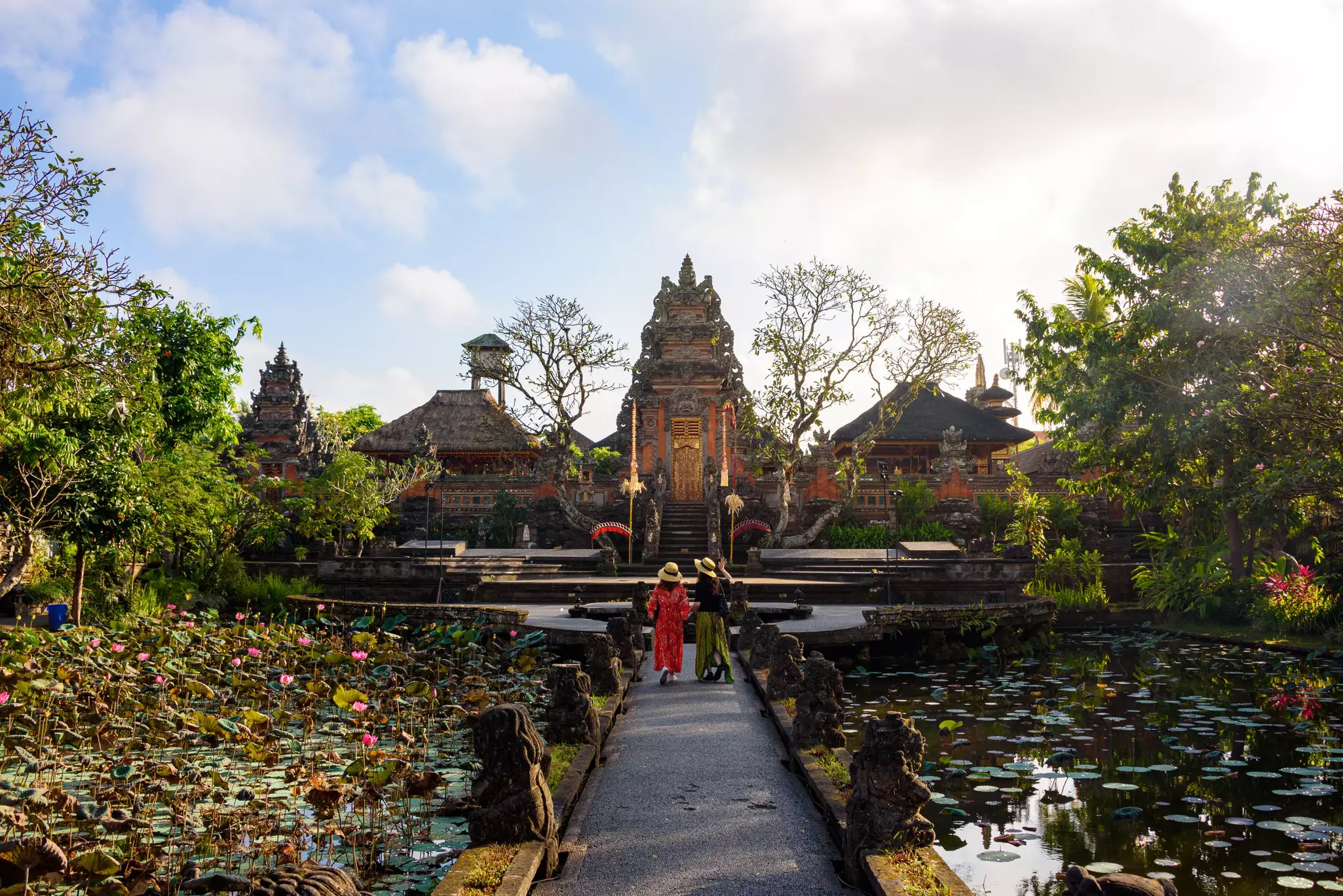 Two people walk on a path over a pond with water lilies toward a Balinese temple at dawn.