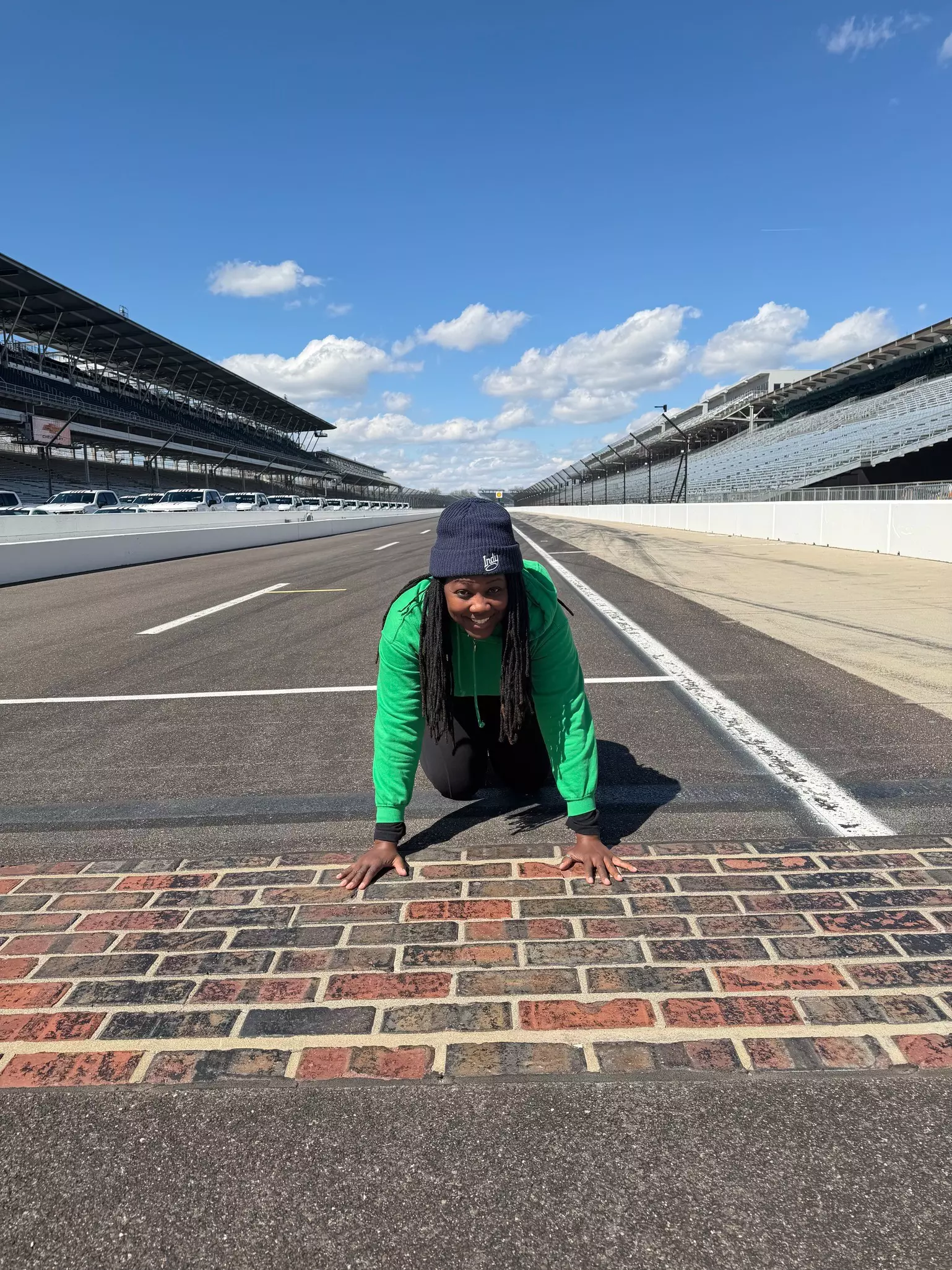A smiling Black woman with locks, wearing a green shirt and and dark pants and hat poses on a racetrack
