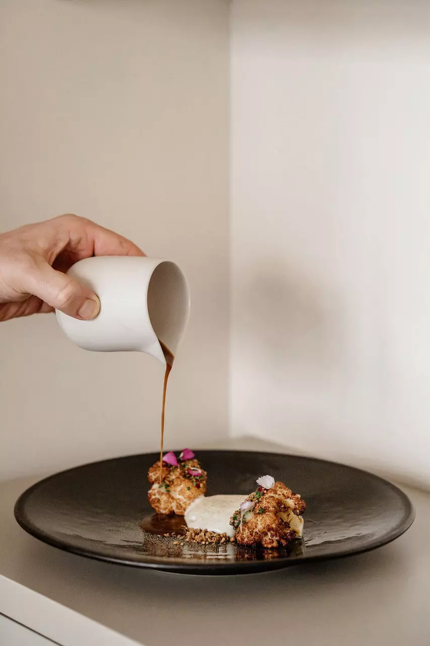 A hand pours sauce from a tiny white jug onto a delicately presented plate of vegan food.