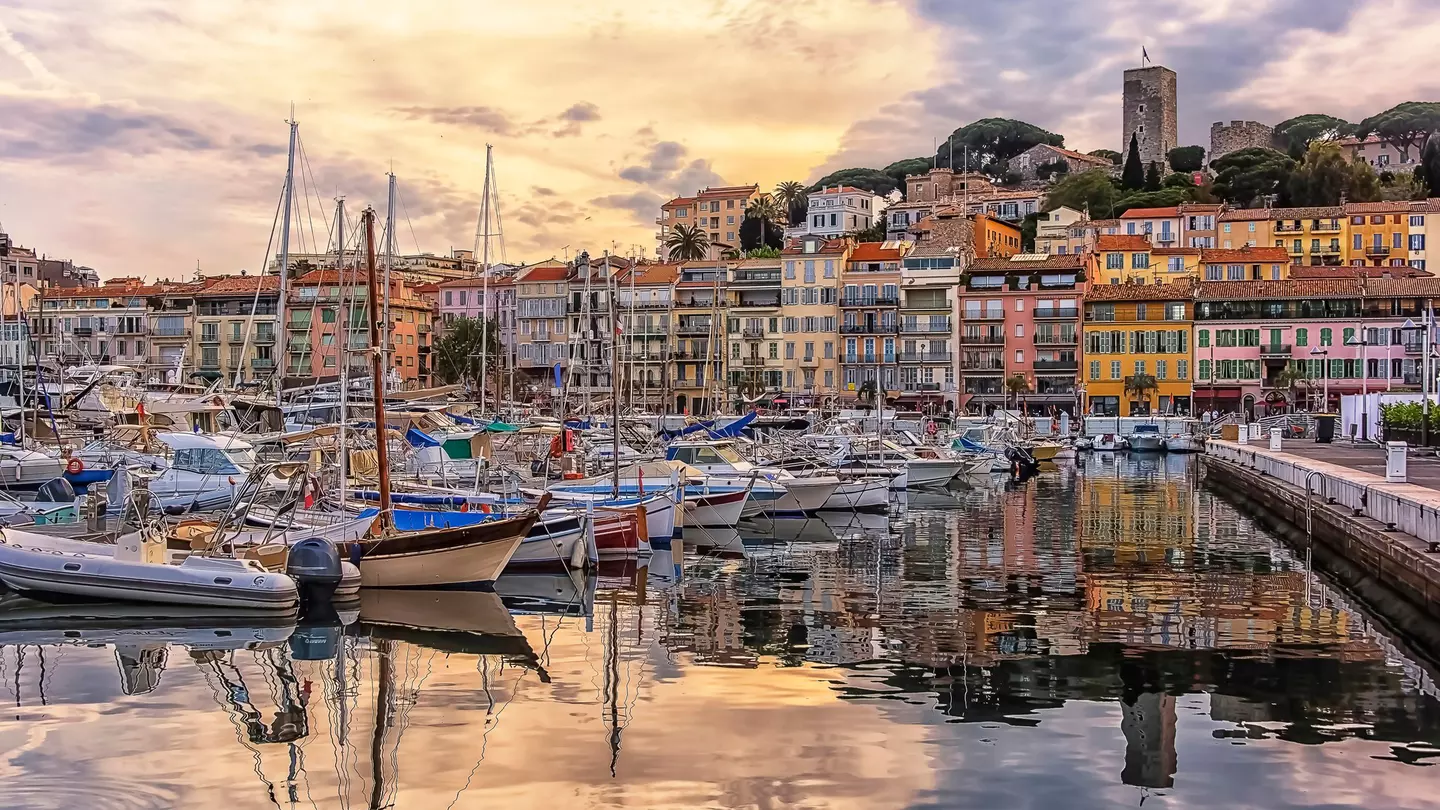 Boats are moored in a port at sunset. Their outlines are reflected in the water. A line of colorful buildings lines the shore, and a castle sits on a hill that rises behind them.