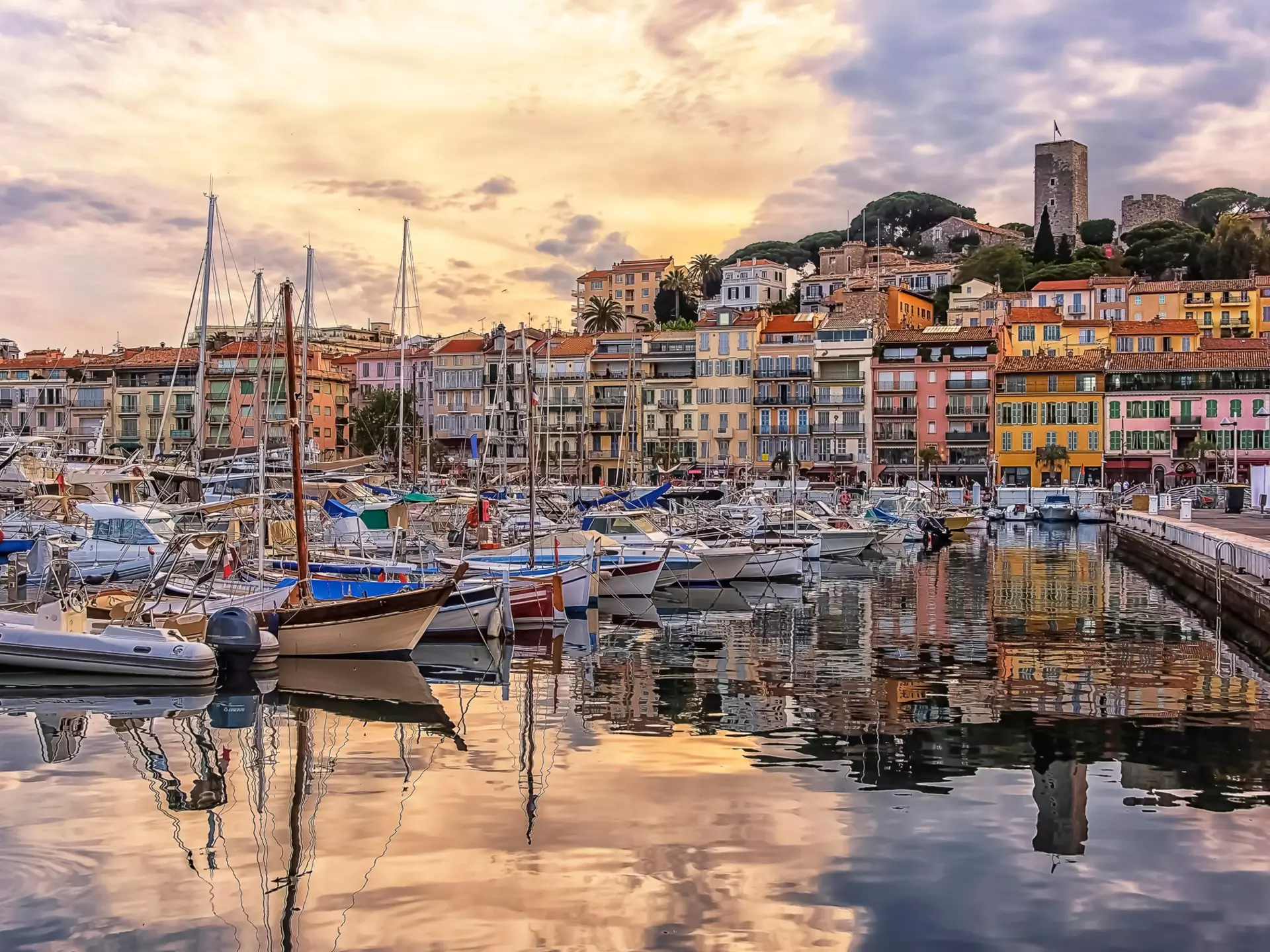 Boats are moored in a port at sunset. Their outlines are reflected in the water. A line of colorful buildings lines the shore, and a castle sits on a hill that rises behind them.