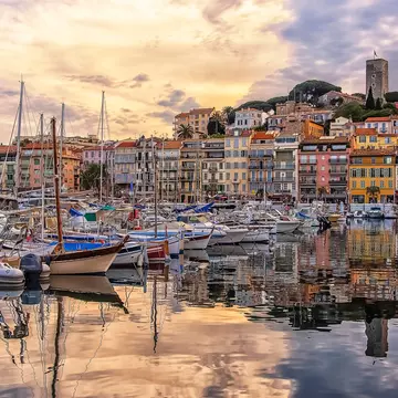 Boats are moored in a port at sunset. Their outlines are reflected in the water. A line of colorful buildings lines the shore, and a castle sits on a hill that rises behind them.
