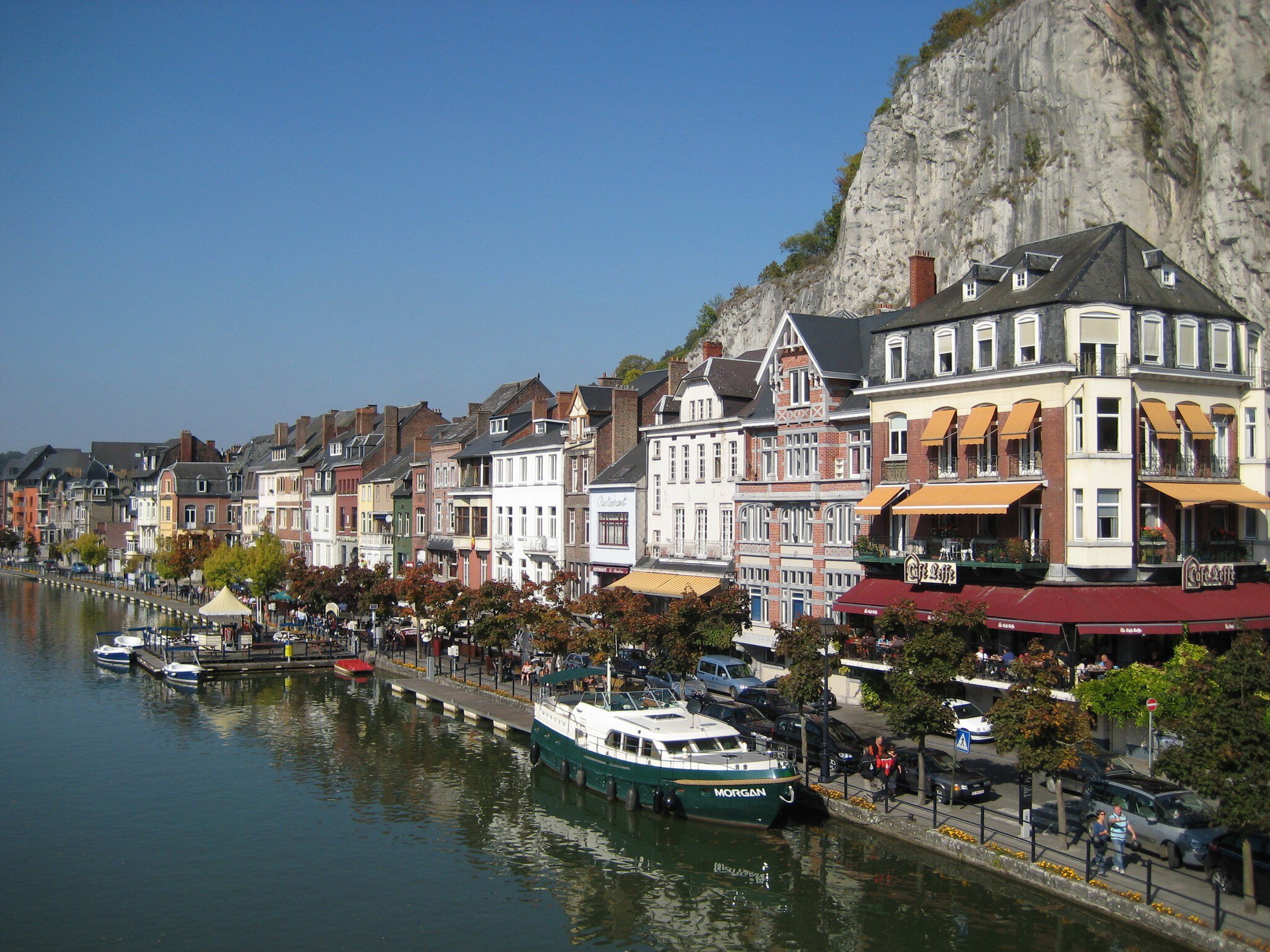 Colorful riverside buildings housing restaurant and bars line the river in Dinant, with boats moored along the waterfront.