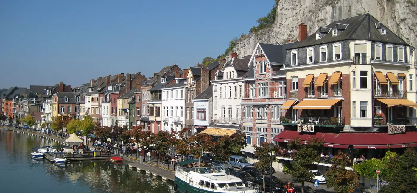 Colorful riverside buildings housing restaurant and bars line the river in Dinant, with boats moored along the waterfront.