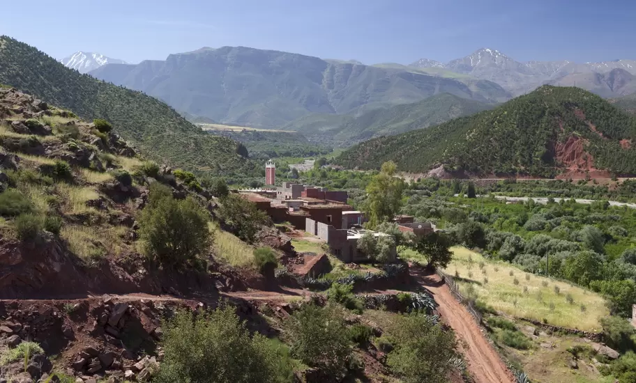 Red-earth village with a minaret in the green Ourika Valley, below snow-capped High Atlas peaks, a dirt track winding in