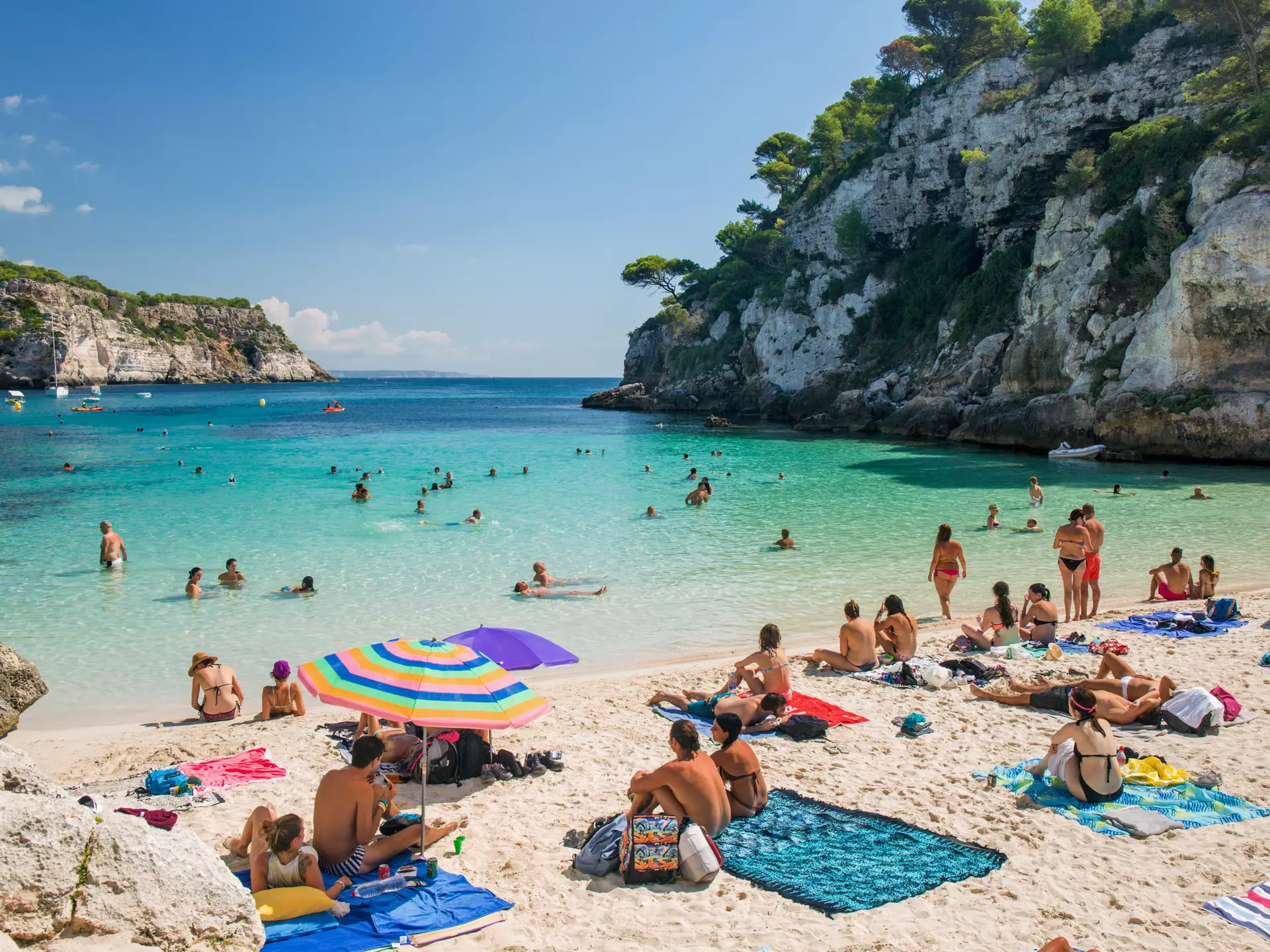 A beach in Menorca, Spain with people sitting on blankets and swimming in the water. 