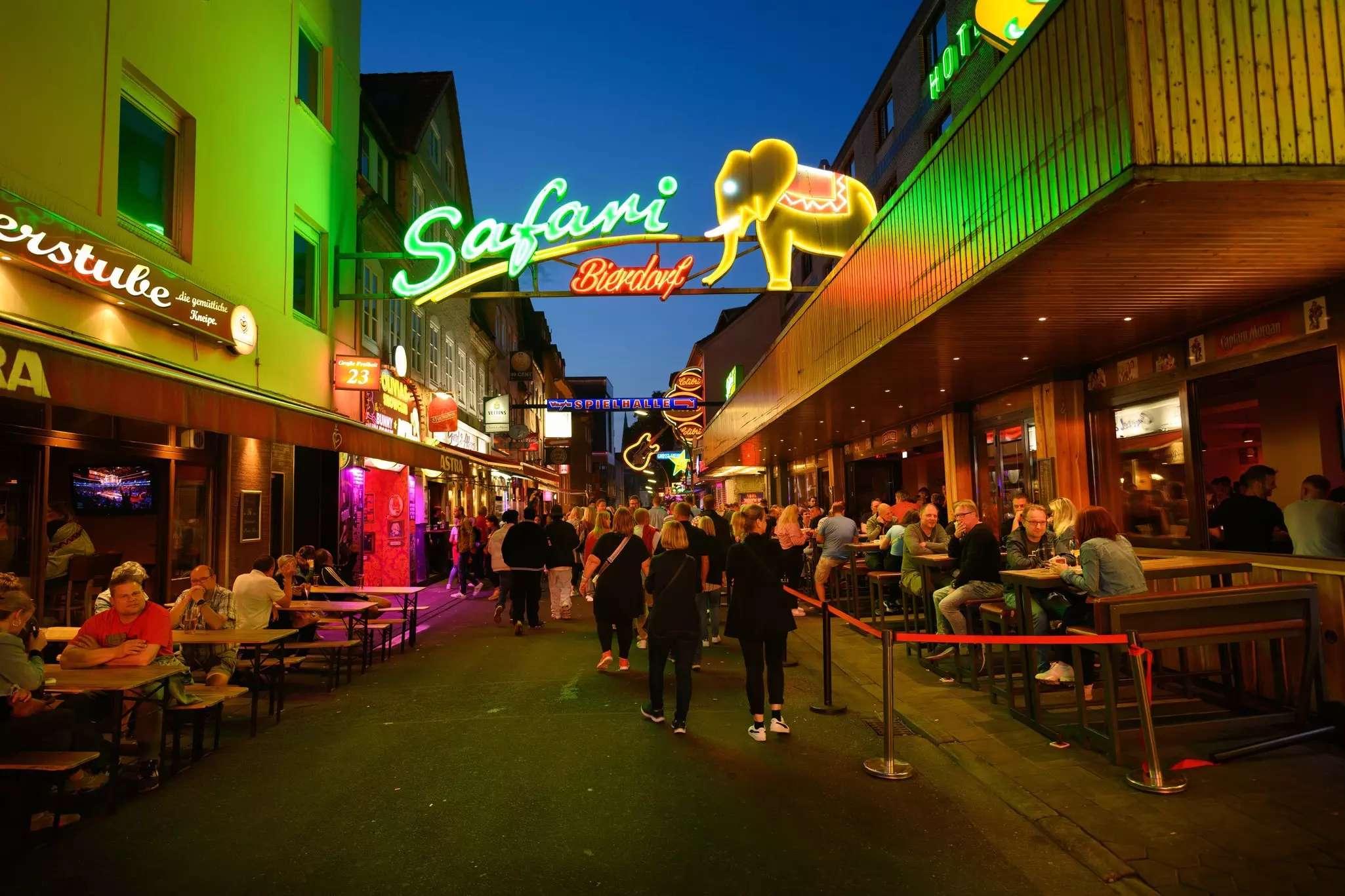 People walking down a narrow pedestiran street with pubs on either side and a neon sign with "Safari Bierdorf" and an elephant overhead at night.