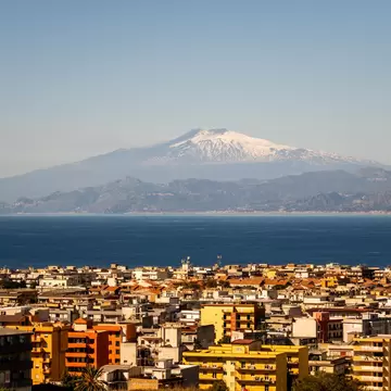 Apartment blocks of Reggio Calabria in the foreground, with blue sea and a snow-topped Mount Etna on the far shore.