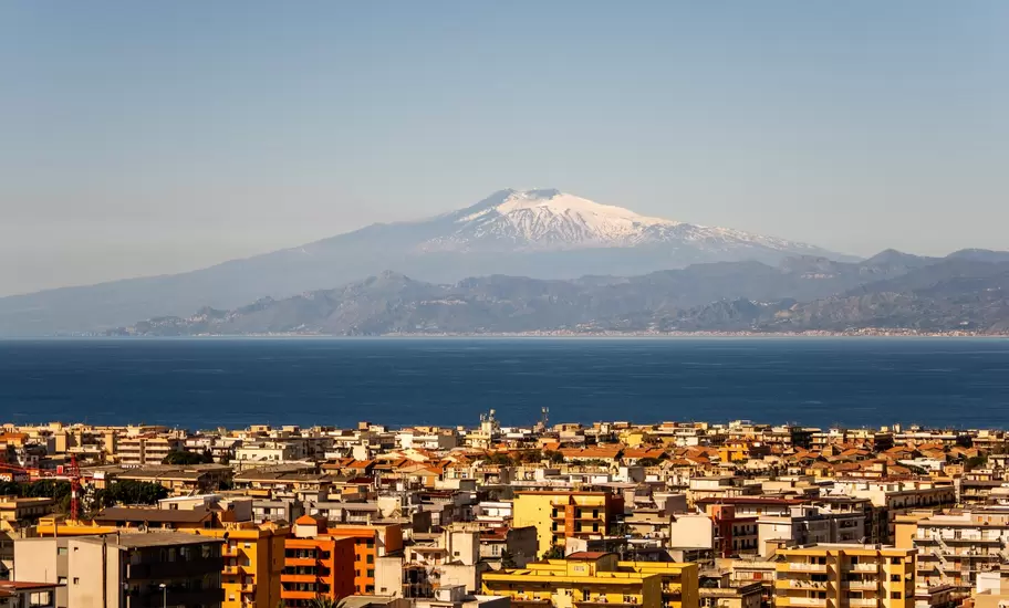 Apartment blocks of Reggio Calabria in the foreground, with blue sea and a snow-topped Mount Etna on the far shore.