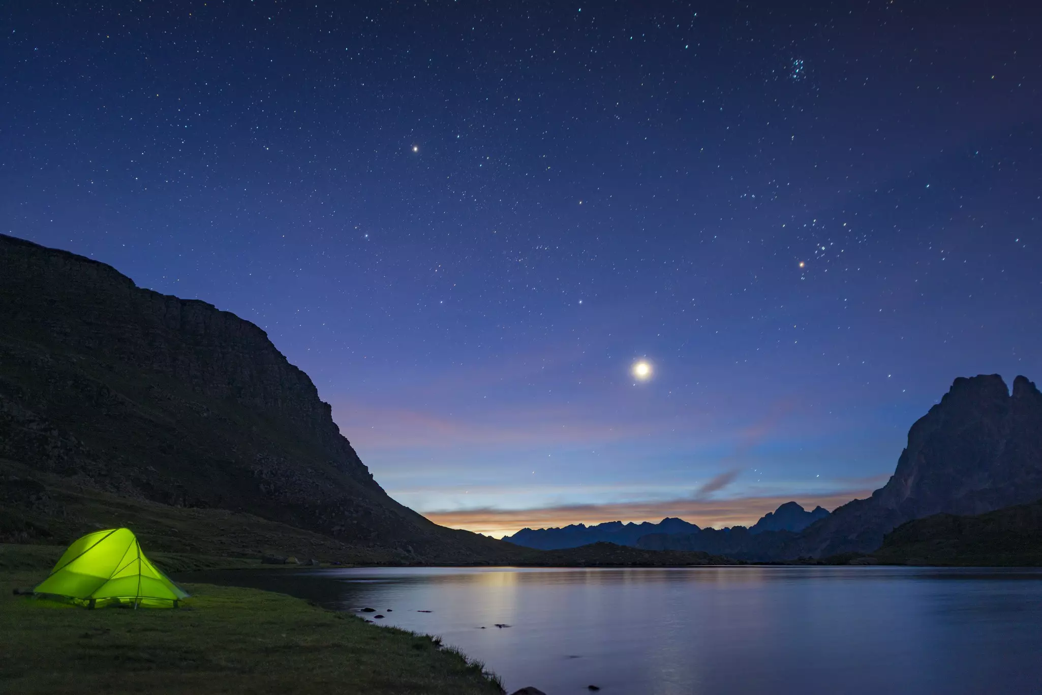 A tent glows with green ligh on the side of a lake, with a sky full of stars above.
