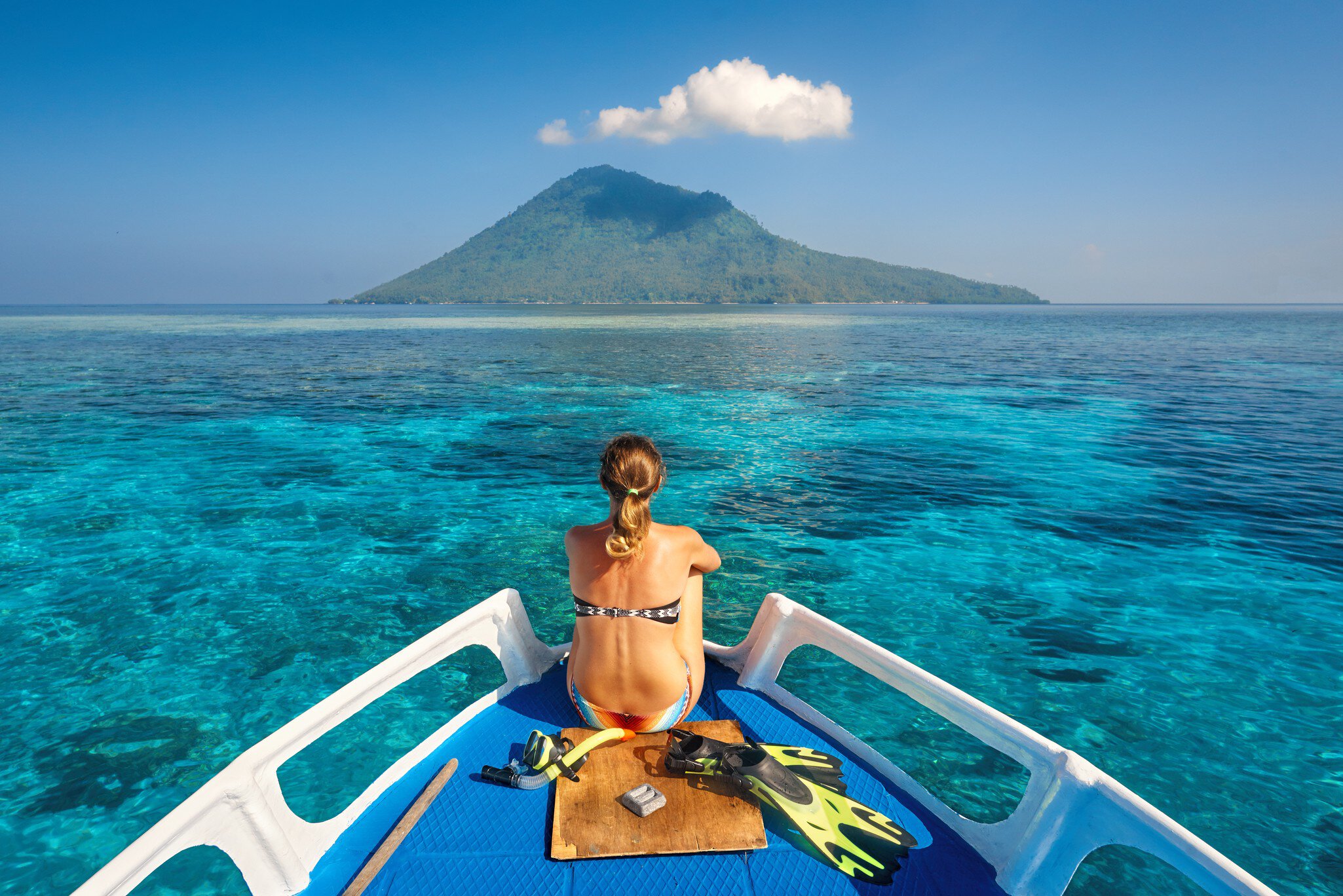 Young woman in swimsuit sit on boat with a mask and flippers looking to a clean sea and volcano Manado Tua. North Sulawesi, Indonesia.