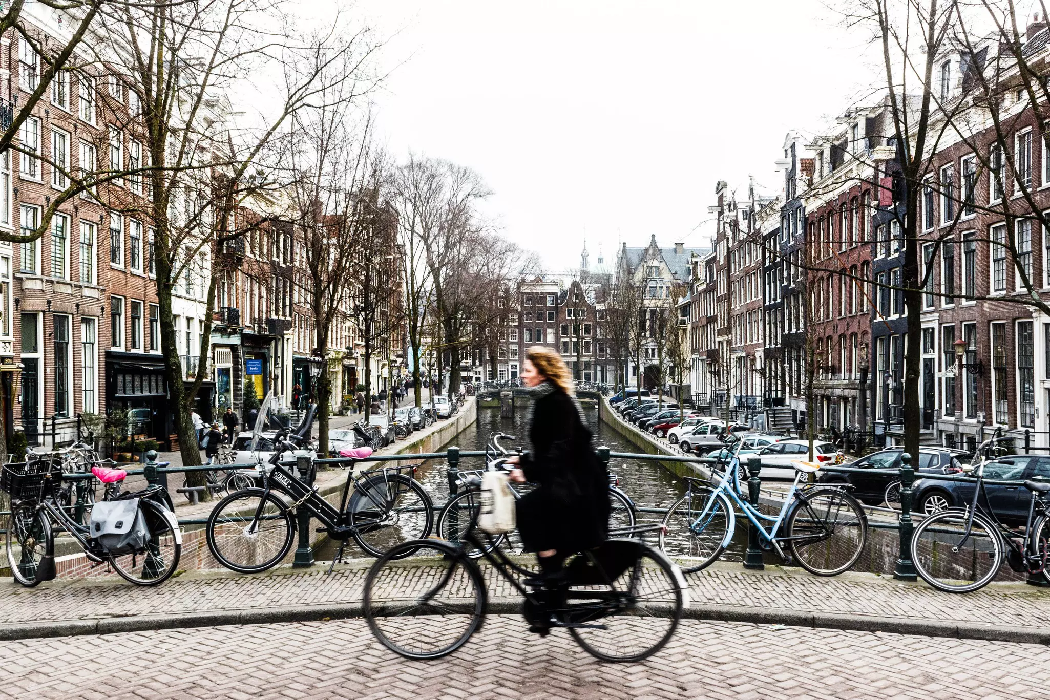 Woman in black out-of-focus as she bicycles on a brick bridge over a canal with otehr bikes locked to the bridge and parked cars and buildings in the distance.