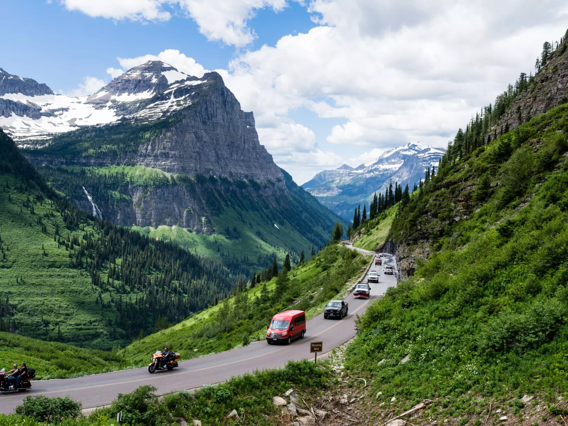 A line of cars drives along green grass and a mountain road