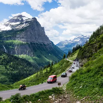 A line of cars drives along green grass and a mountain road