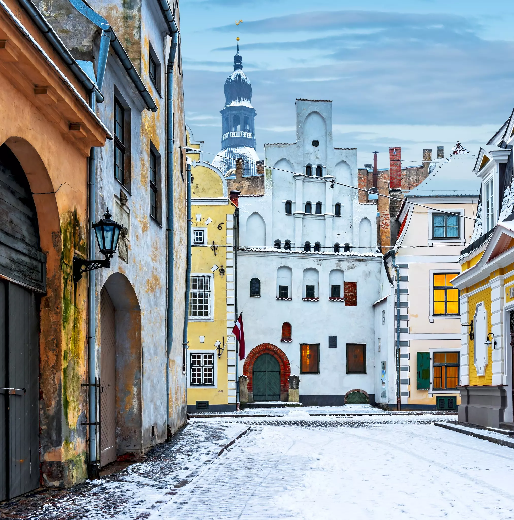 A snow-covered cobbled street leads to a medieval row of buildings, with the central one built with a distinct sloping angle to its roof and windows.