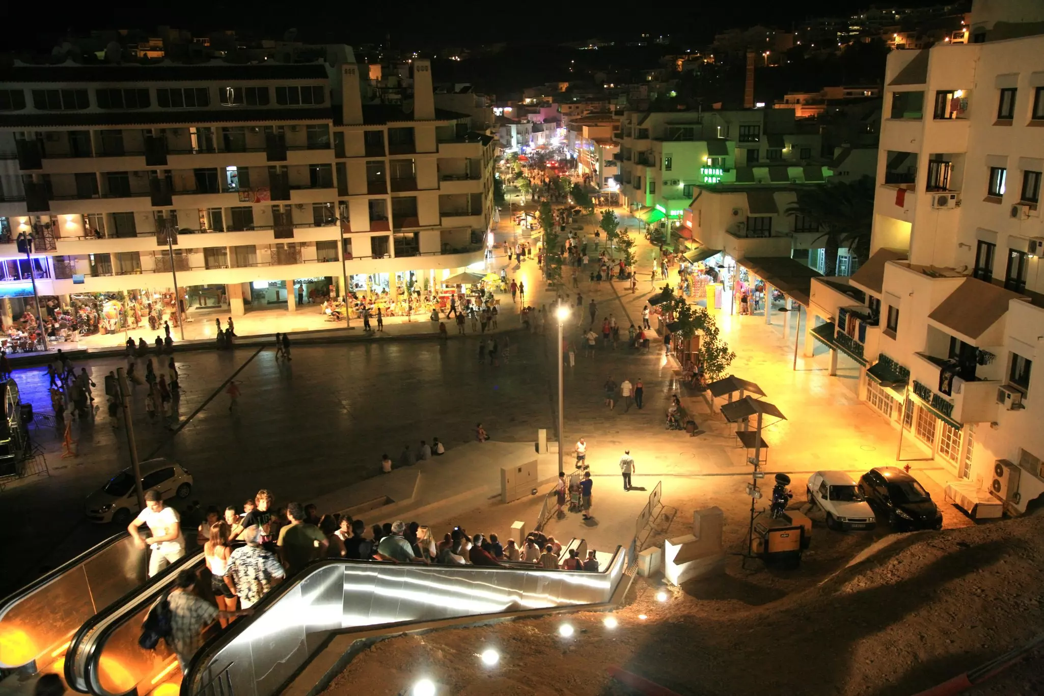 People ride a public escalator leading up from a city square in the evening. Bars and restaurants are seen on a street in the distance.