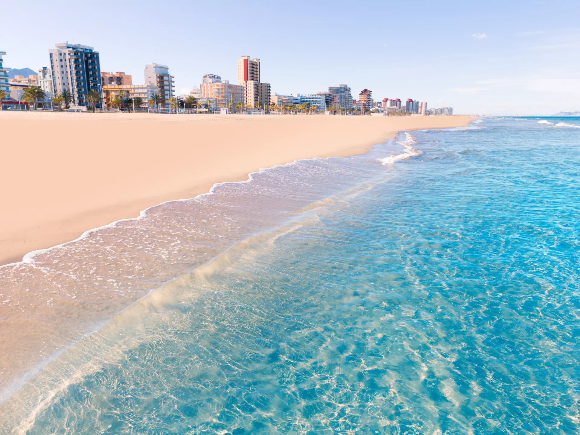 The clear, blue wash of the Mediterranean Sea cuts diagonally against the soft, pinkish sands of Playa de Gandia in Valencia, Spain with several tower blocks rising up in the background