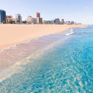 The clear, blue wash of the Mediterranean Sea cuts diagonally against the soft, pinkish sands of Playa de Gandia in Valencia, Spain with several tower blocks rising up in the background