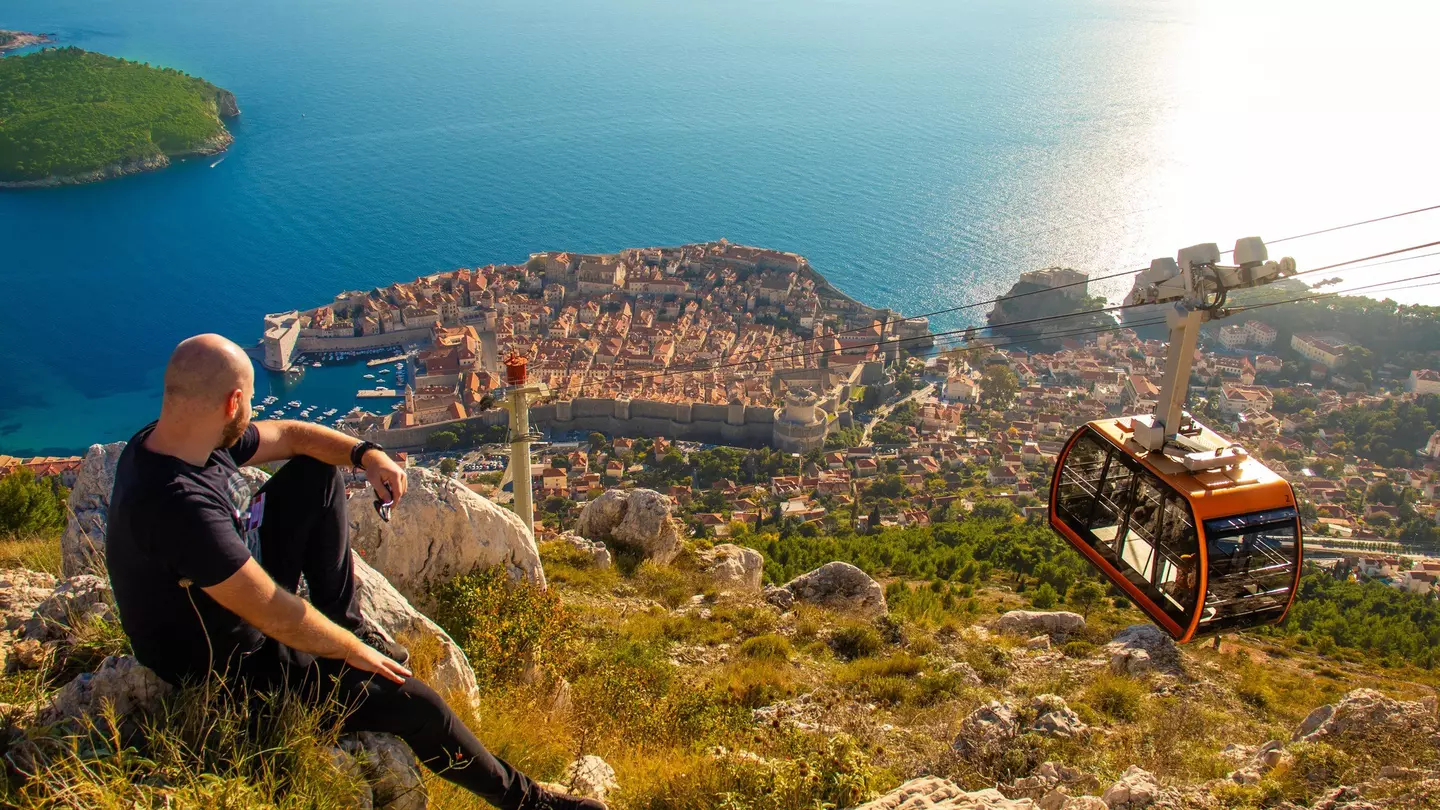 A man sits on a rock looking down at a coastal city as a cable car passes by