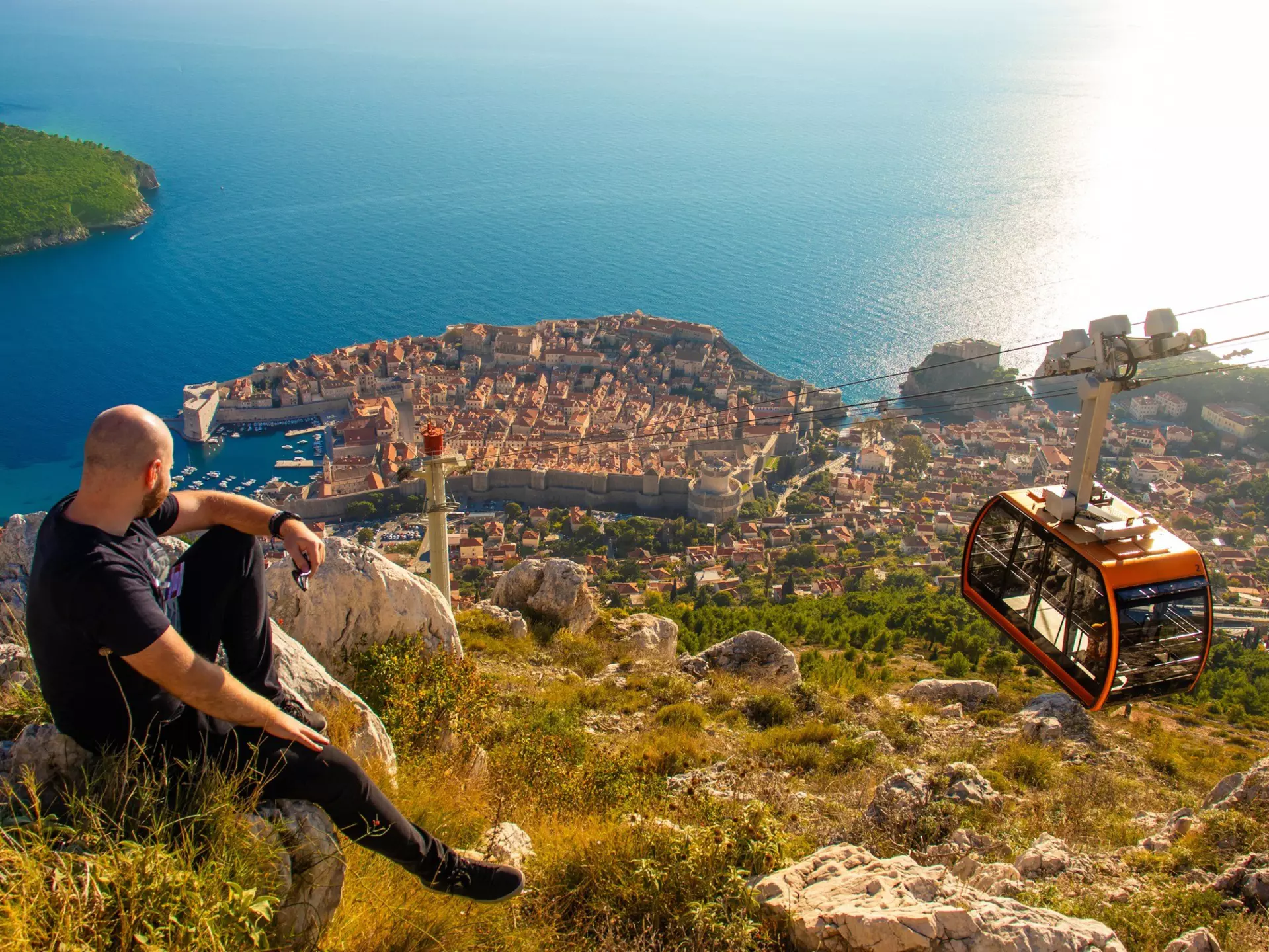 A man sits on a rock looking down at a coastal city as a cable car passes by