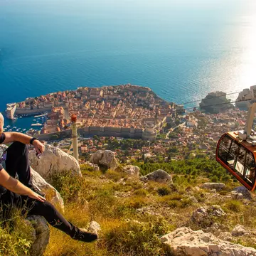 A man sits on a rock looking down at a coastal city as a cable car passes by