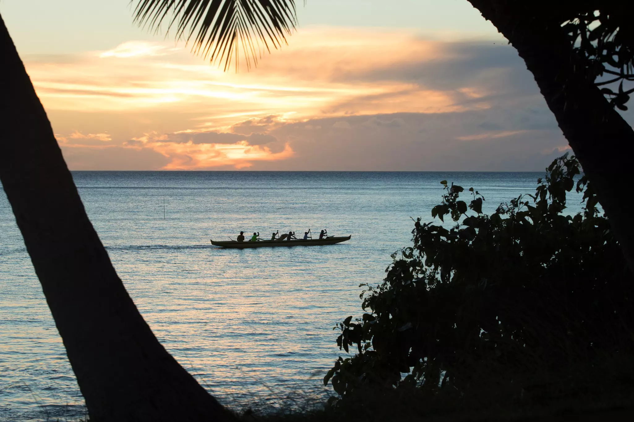 Seven people in silhouette as they paddle a long canoe through the ocean at sunset.