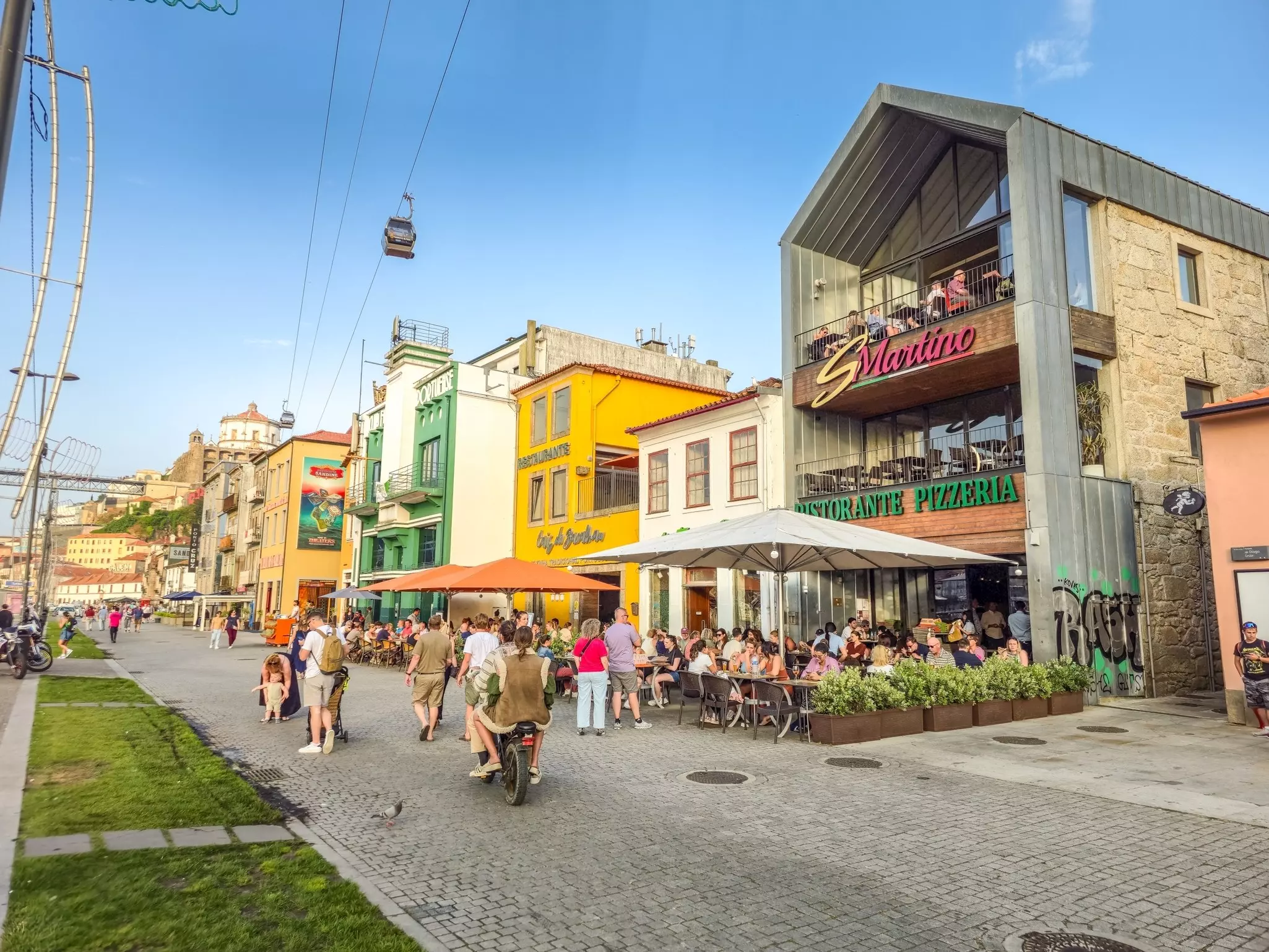 People walk along a cobbled street in front of restaurant terraces. A cable car passes overhead.