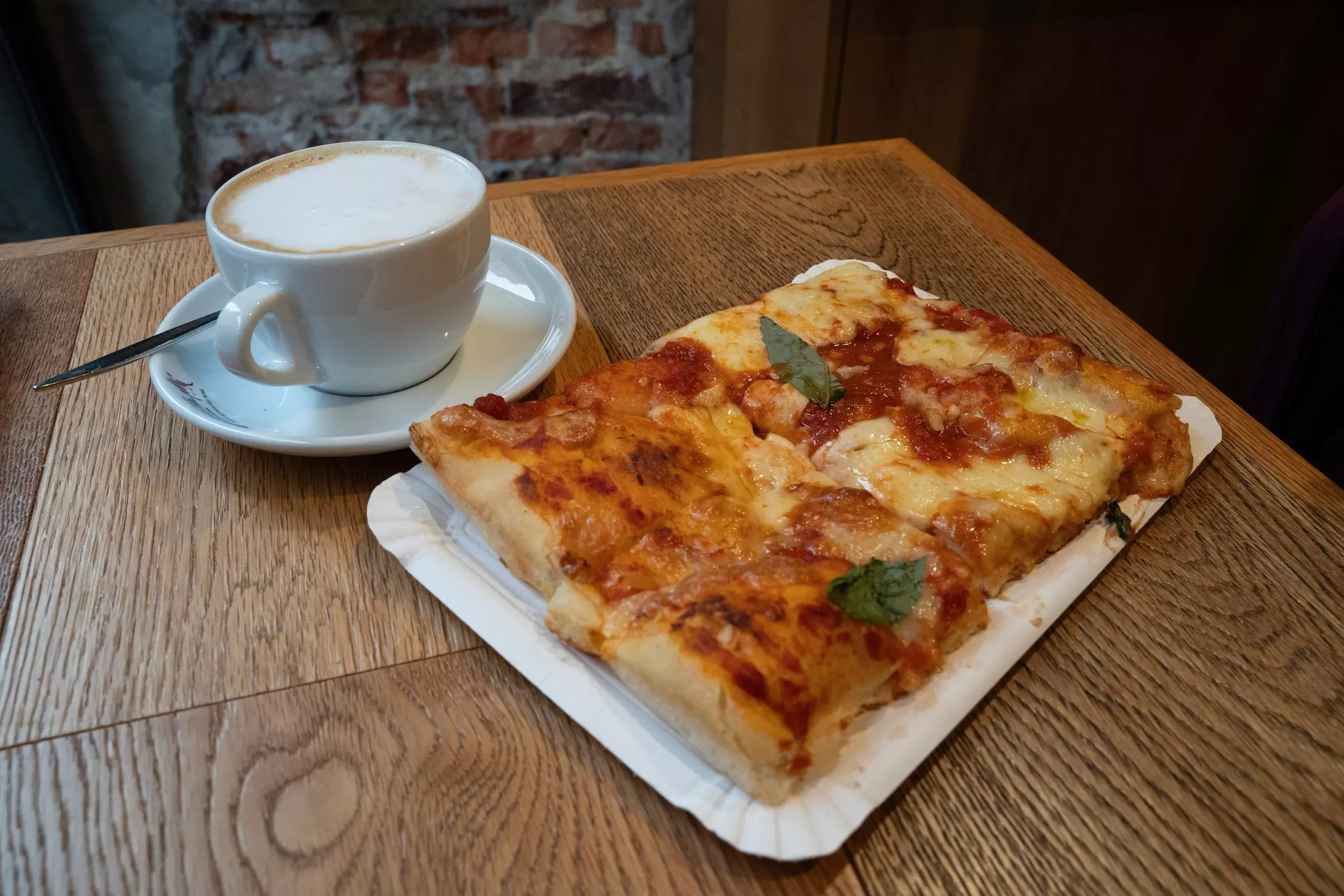 A cappuccino in a white cup sits next to a large piece of focaccia on a wooden tabletop.