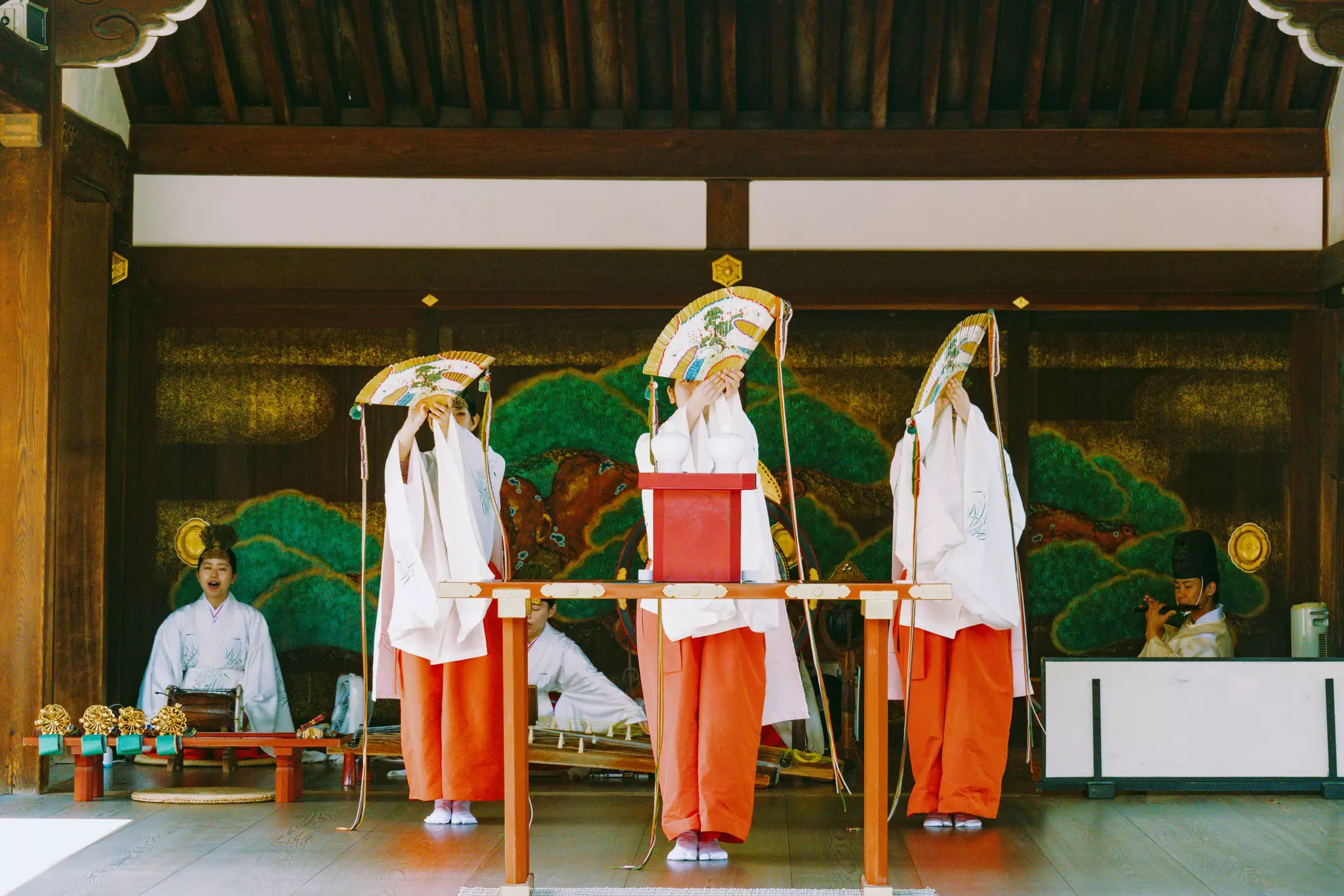 Three women in white tops and orange pants perform with fans on a stage.