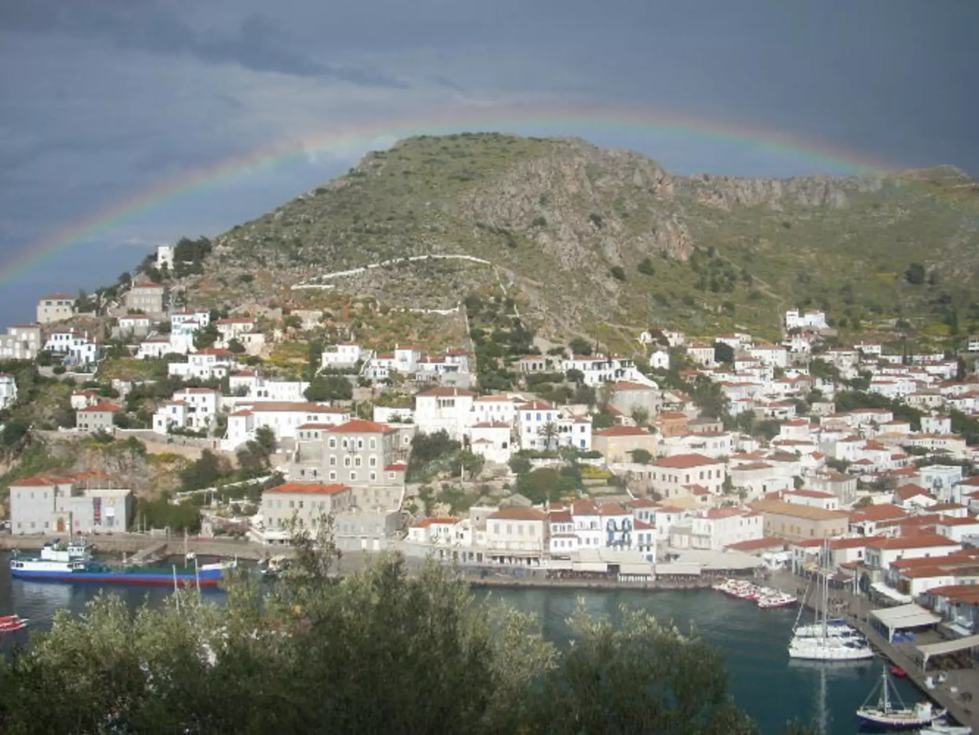 Many white buildings with red roofs line the bottom of a hill. There's a small harbor in the foreground and a rainbow over the hill