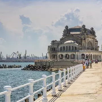 A white casino building on the Black Sea shore, people on a promenade lined by a balustrade and harbour cranes beyond.