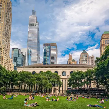 A section of green space among tall skyscrapers. People are relaxing on the grass on a sunny day.