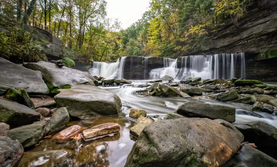 Wide curtain waterfall spilling over a dark shale ledge into a boulder-strewn creek in Ohio, ringed by early autumn woodland